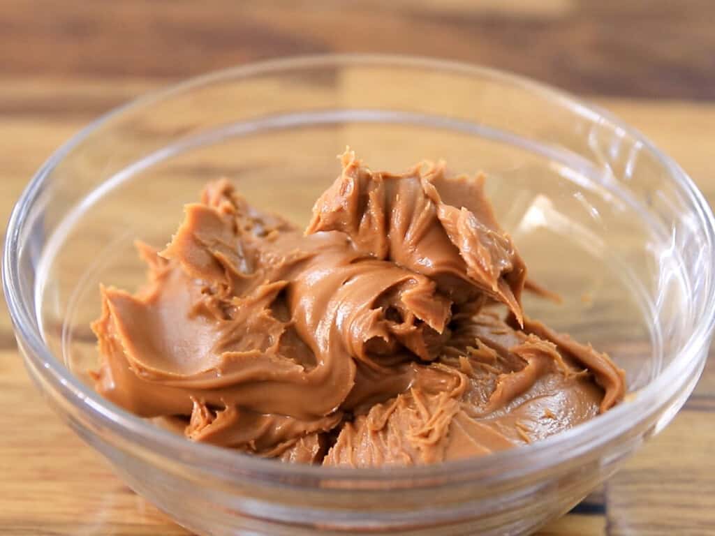 A clear glass bowl filled with a generous dollop of creamy Biscoff cookie butter sits on a wooden surface.