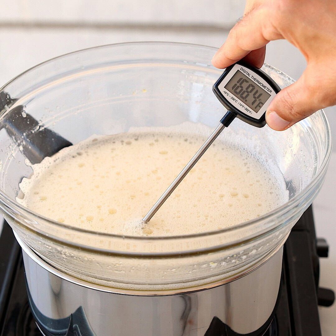 A hand holds a digital thermometer in a glass bowl of frothy liquid set over a pot of boiling water on a stove, checking the temperature, which reads 168.4°F.