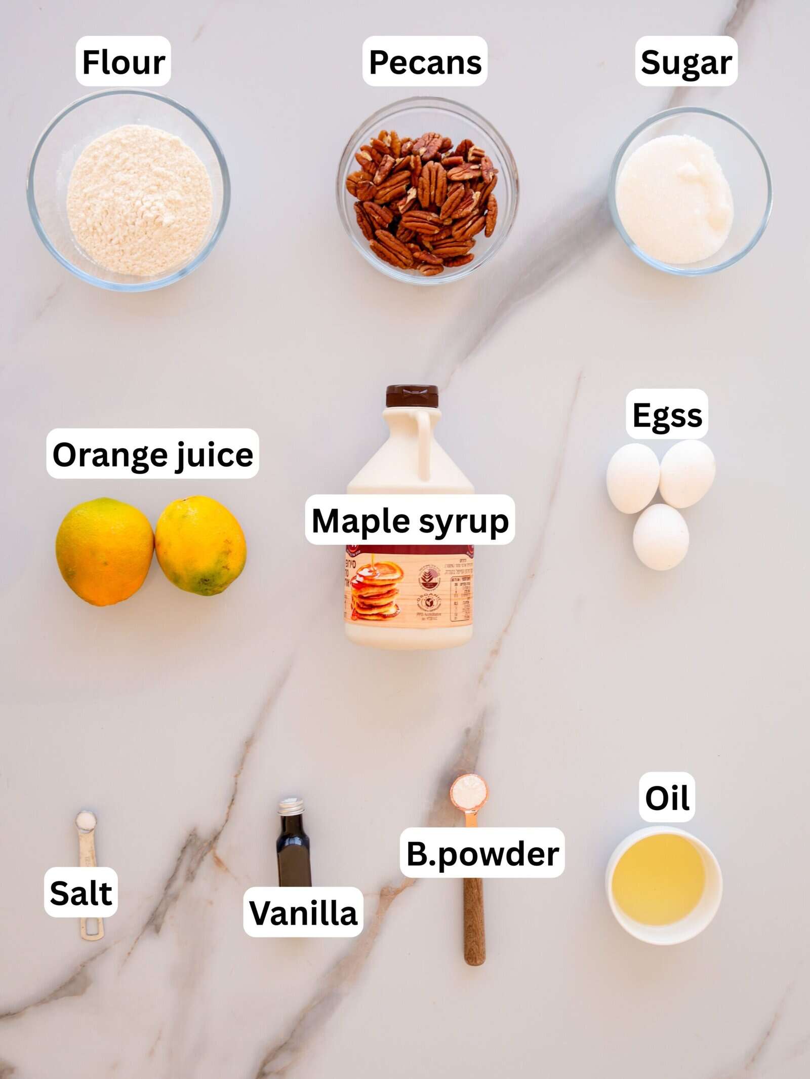 A top view of baking ingredients on a marble surface, labeled: flour, pecans, sugar, orange juice, maple syrup, eggs, oil, salt, vanilla, and baking powder, each in a separate bowl or container.