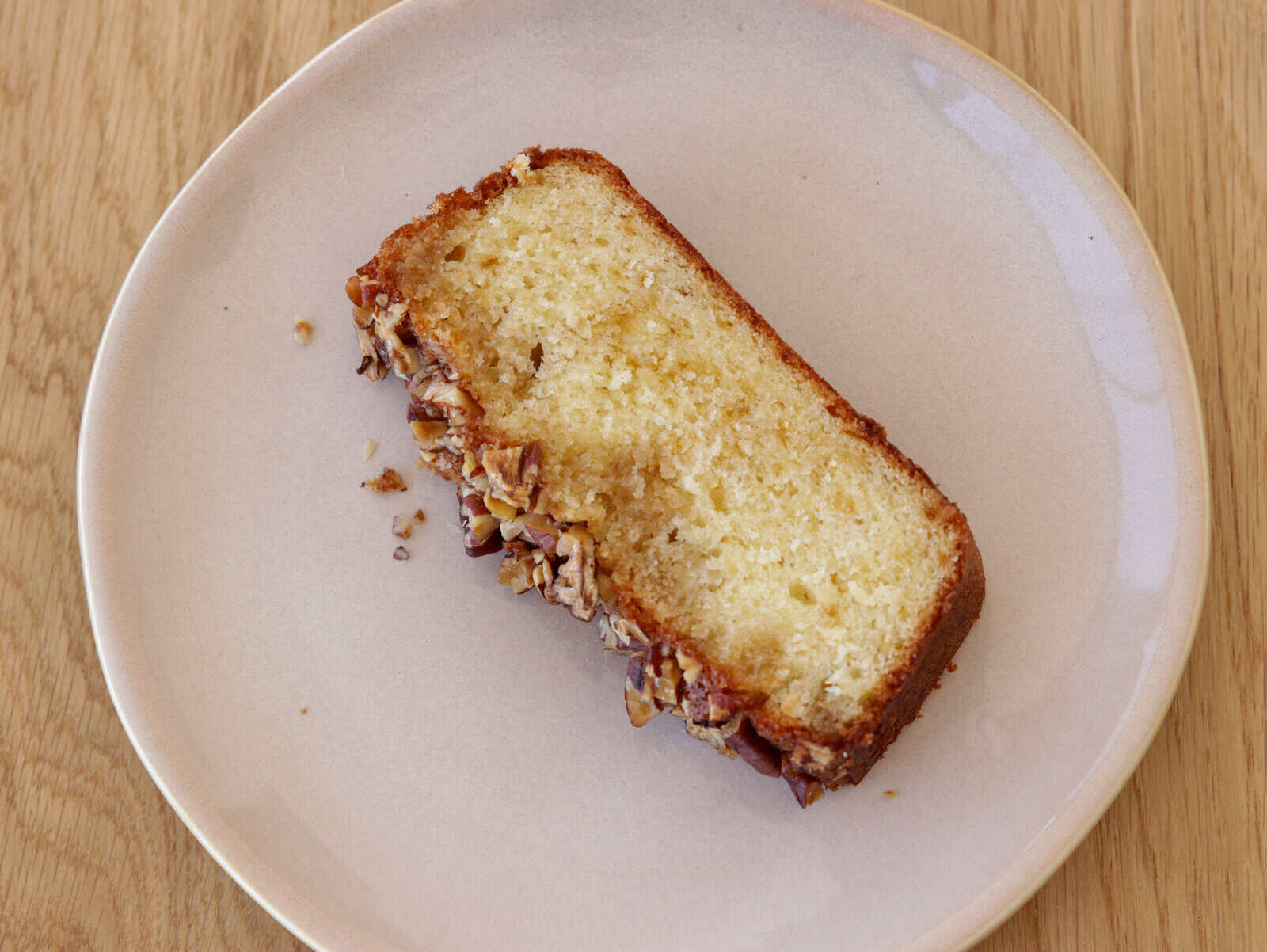 A single slice of nut-topped maple syrup pound cake sits on a light pink plate, which rests on a wooden surface.