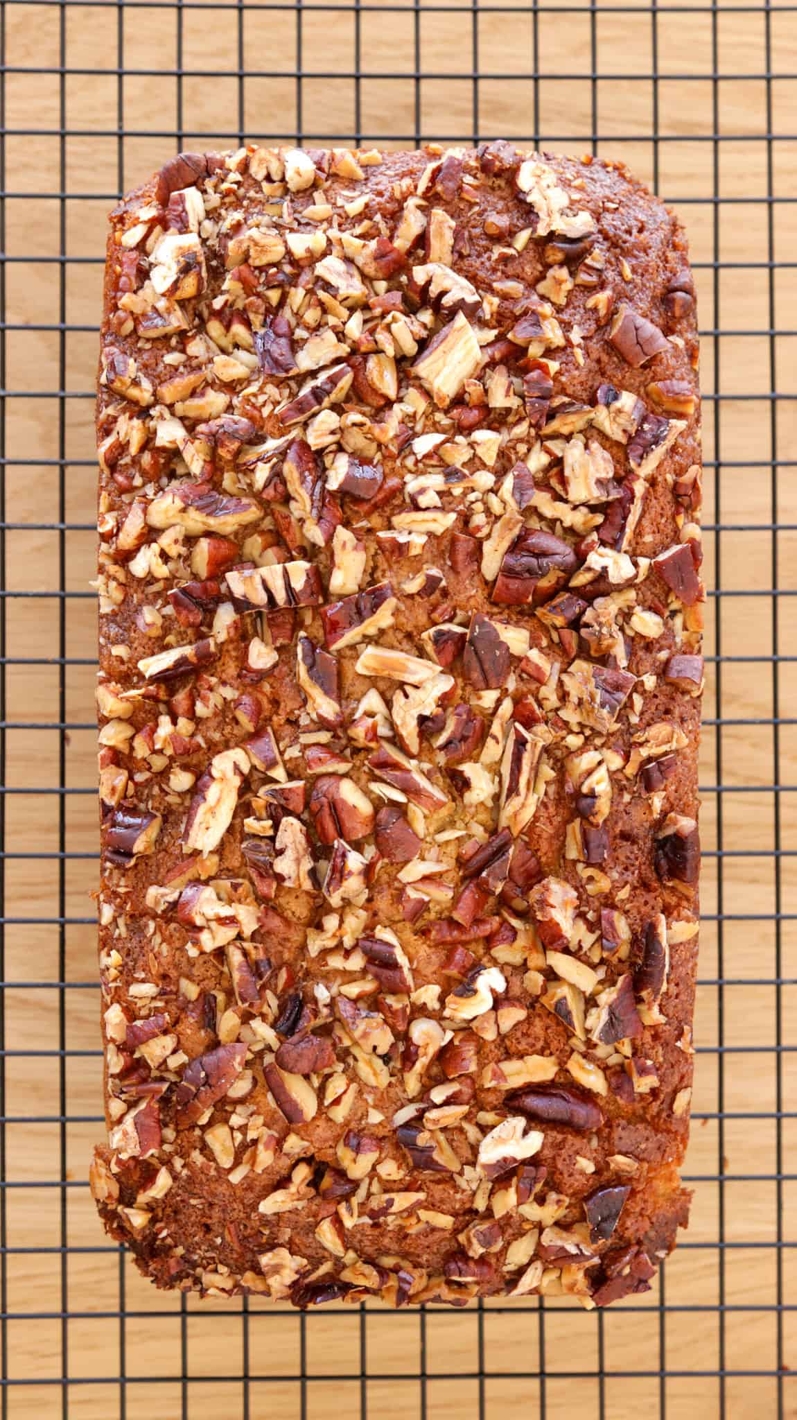 A loaf of nut-topped maple cake, sits on a black wire cooling rack over a wooden surface. The golden-brown bread is covered with chopped pecans.