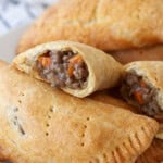 A plate of golden-brown hand meat pies, some whole and one cut open, revealing a savory filling of ground meat and diced vegetables. A white and blue towel is visible in the background.