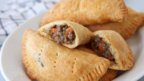A plate of golden-brown hand meat pies, some whole and one cut open, revealing a savory filling of ground meat and diced vegetables. A white and blue towel is visible in the background.