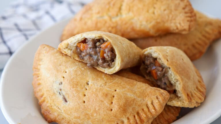 A plate of golden-brown hand meat pies, some whole and one cut open, revealing a savory filling of ground meat and diced vegetables. A white and blue towel is visible in the background.