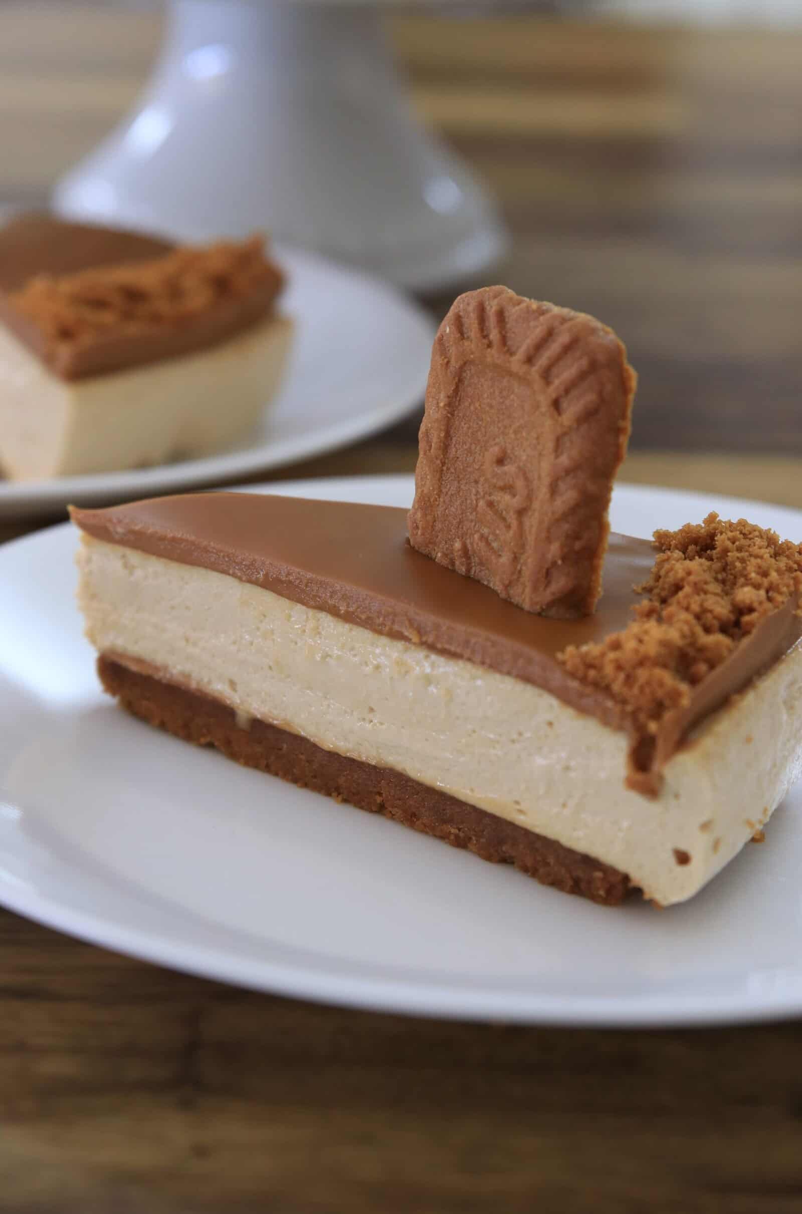 A slice of a no-bake bisocff cheesecake with a biscuit base, creamy filling, caramel-colored topping, and a whole biscuit and crumbs on top, served on a white plate with another slice in the background.