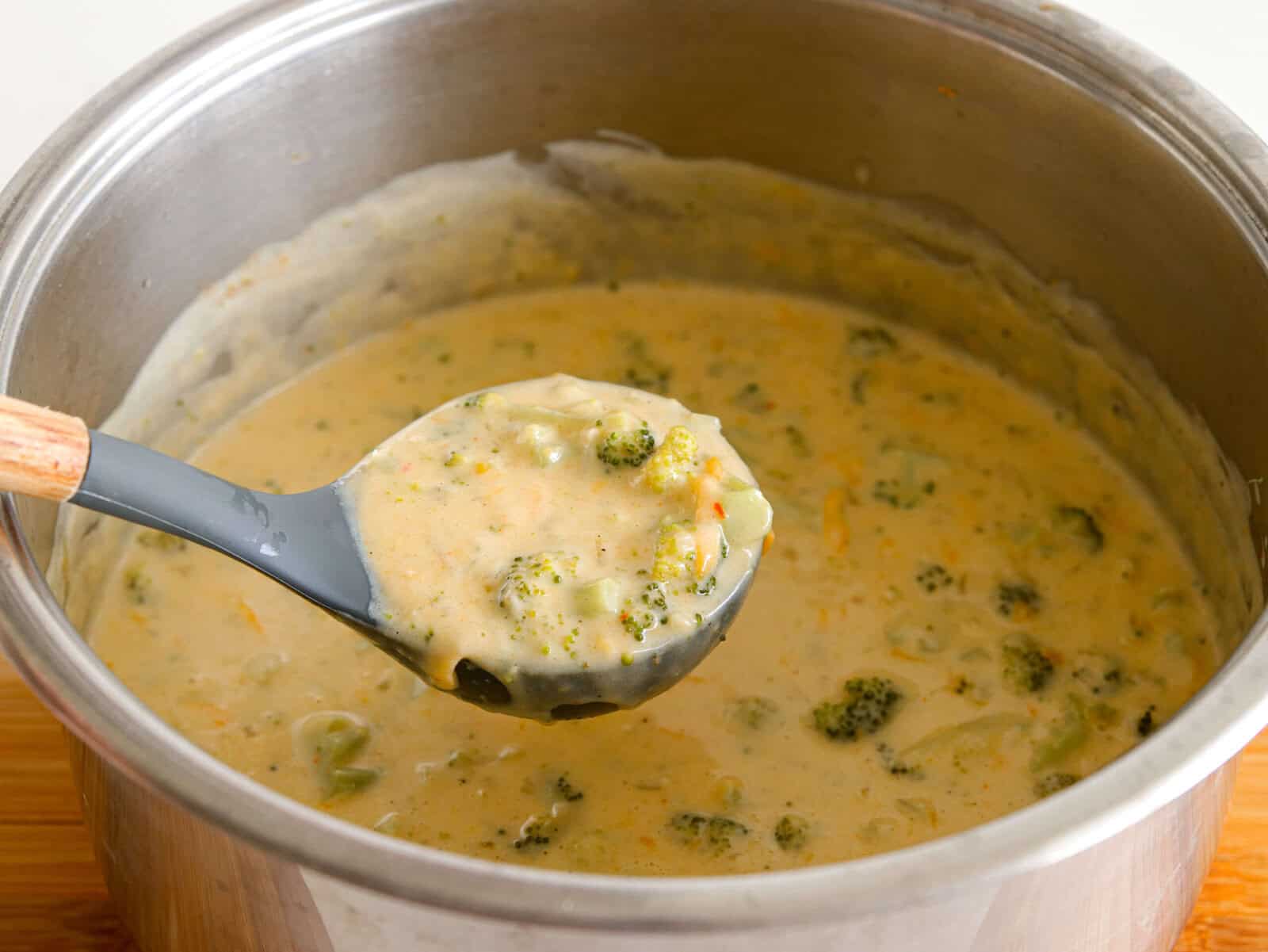 A metal pot filled with creamy broccoli cheddar soup; a ladle lifts a serving showing chunks of broccoli and a thick, cheesy texture.