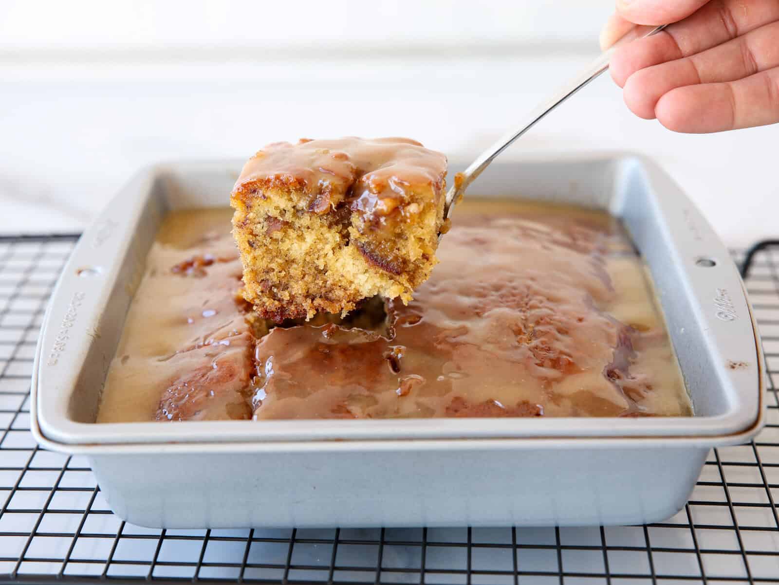 A hand lifts a square piece of glazed sticky date pudding cake from a metal baking pan resting on a cooling rack. The cake is golden brown with a thick, shiny glaze on top.