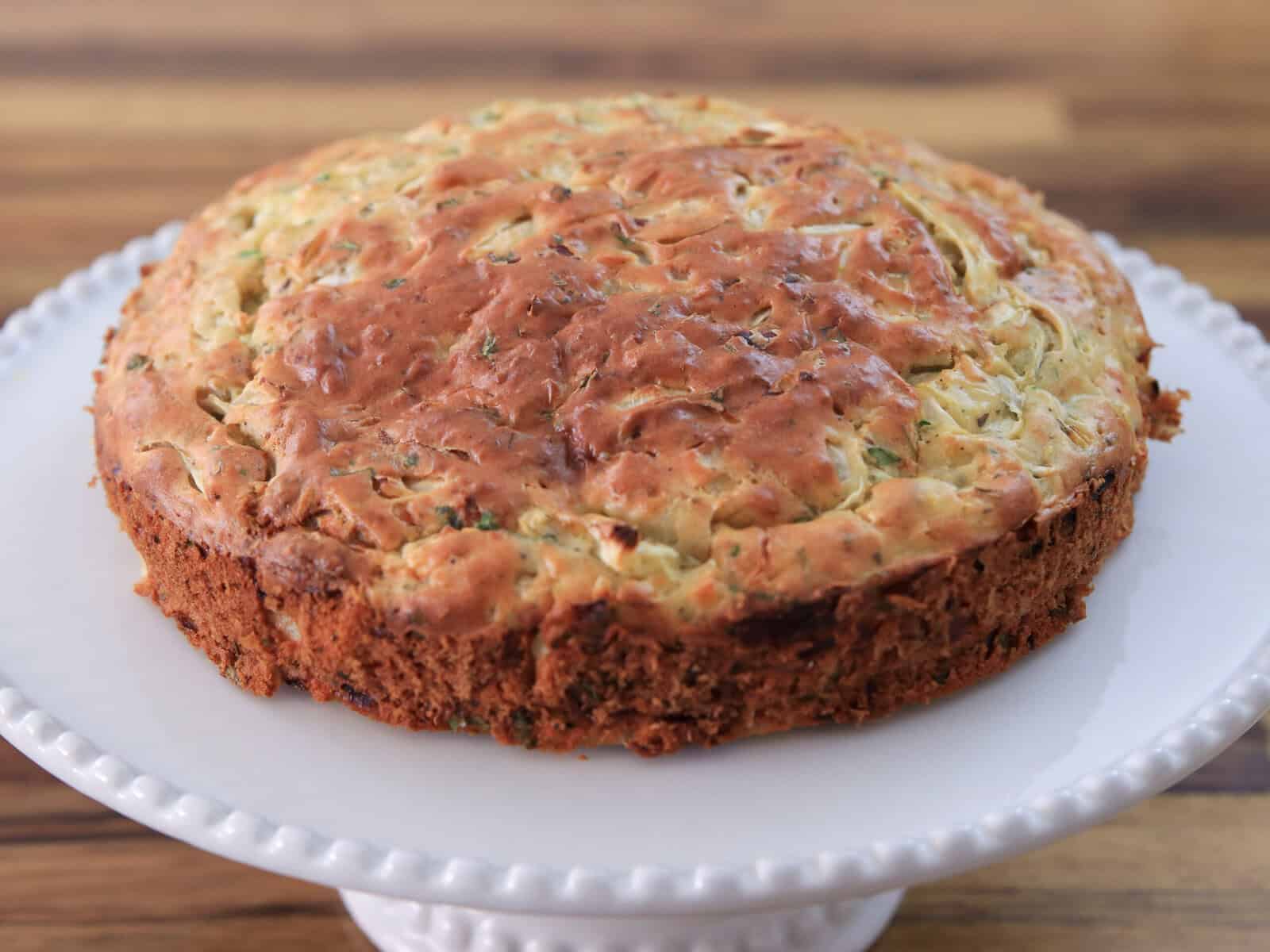 A round, golden-brown savory cabbage cake with chopped herbs is on a white plate. The background is a wooden surface.