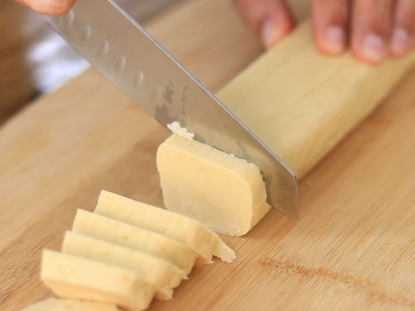 A close-up of a hand using a large knife to slice a log of dough into even pieces on a wooden cutting board.