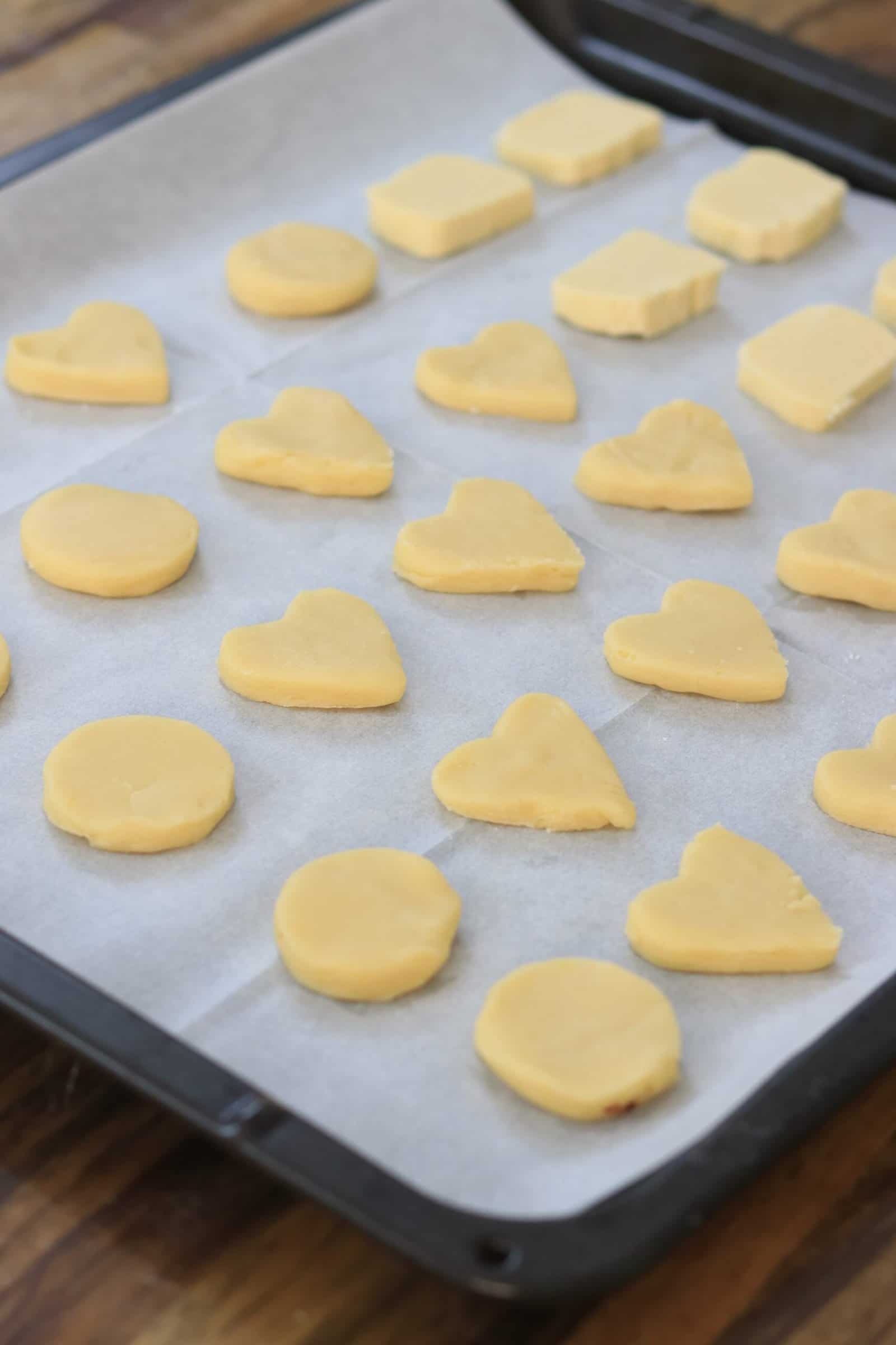 A baking tray lined with parchment paper holds rows of unbaked 3-ingredient butter cookies in heart, round, and rectangular shapes, arranged neatly and ready for baking.