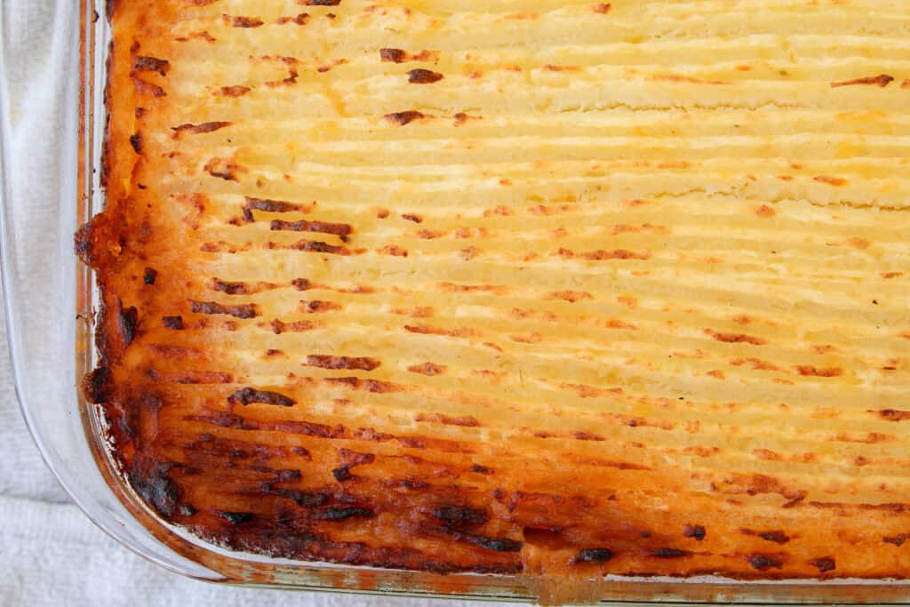 A close-up of a baked shepherd’s pie in a glass dish, showing a golden brown, textured mashed potato topping with slightly crispy edges.