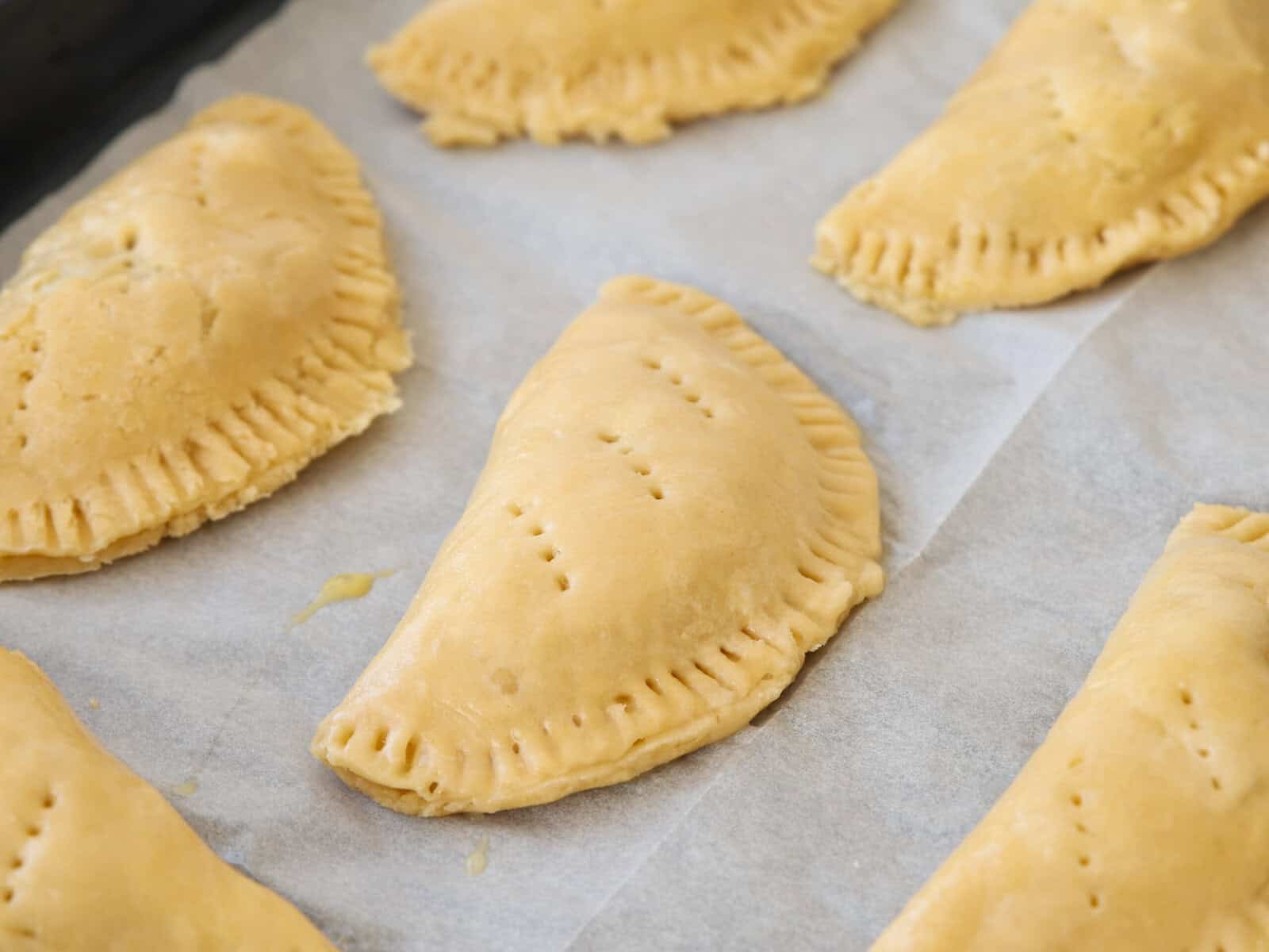 Unbaked Nigerian meat pies arranged on a sheet of parchment paper, with crimped edges and fork holes on top, ready to be baked.