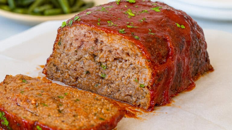 A close-up of a glazed meatloaf with a slice cut off, showing its moist, tender interior. The meatloaf is topped with a shiny red sauce and sprinkled with chopped herbs. Green beans are blurred in the background.