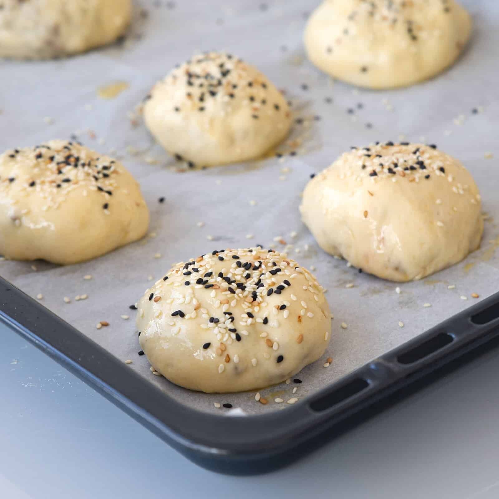 Unbaked dough balls topped with mixed sesame and poppy seeds are arranged on a parchment-lined baking tray, ready to go into the oven.