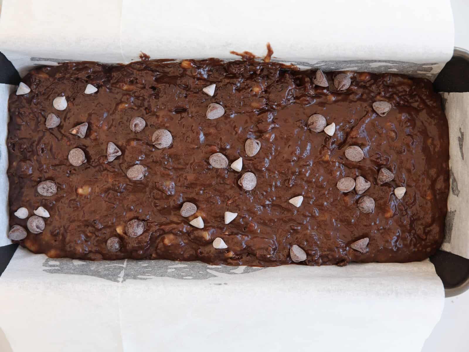 A loaf pan lined with parchment paper filled with unbaked chocolate banana bread batter, topped with scattered chocolate chips.