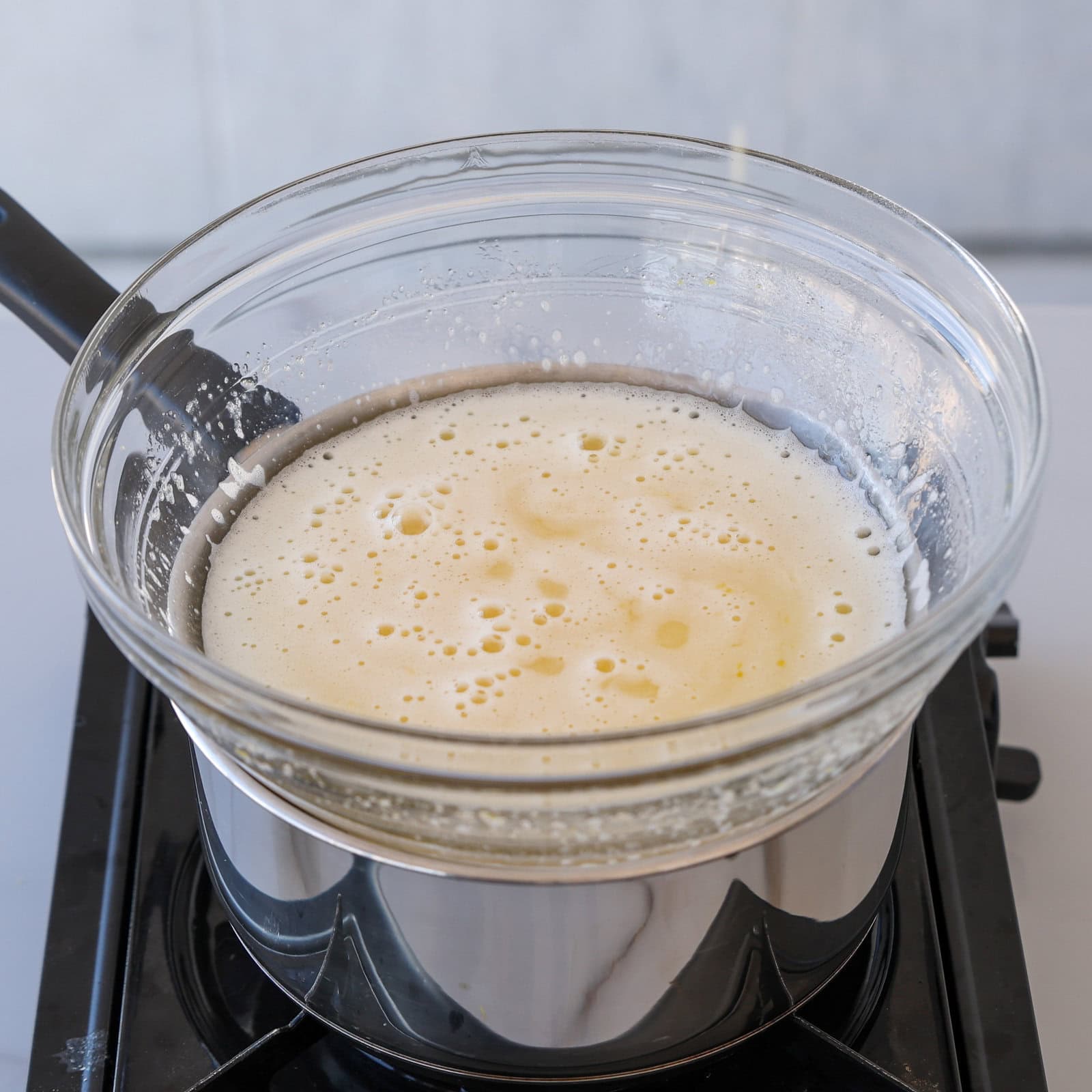 A glass bowl with a frothy, pale yellow mixture sits over a pot of simmering water on a stovetop, forming a double boiler setup.