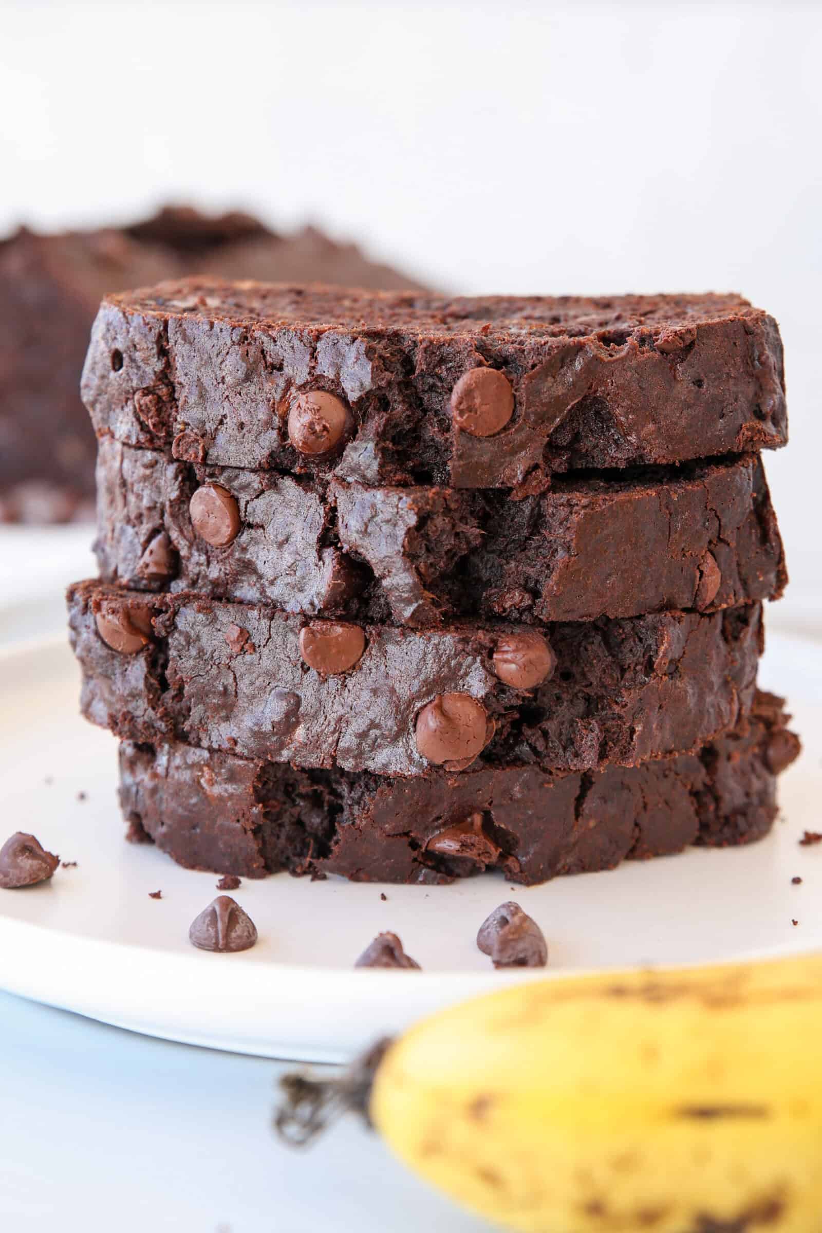 Three thick slices of easy chocolate chip banana bread stacked on a white plate. A few chocolate chips and part of a ripe banana are visible in the foreground.