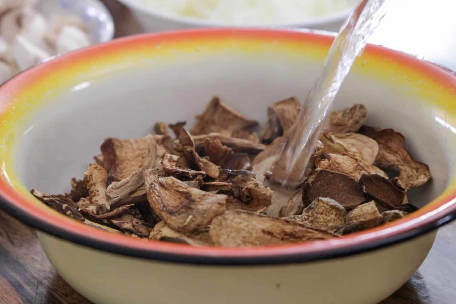 A close-up of dried mushrooms in a bowl as water is being poured over them for rehydration. Other blurred ingredients are visible in the background.