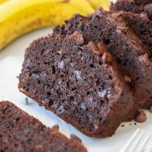 Close-up of sliced chocolate banana bread with chocolate chips, arranged on a white surface, with a few ripe bananas in the background.