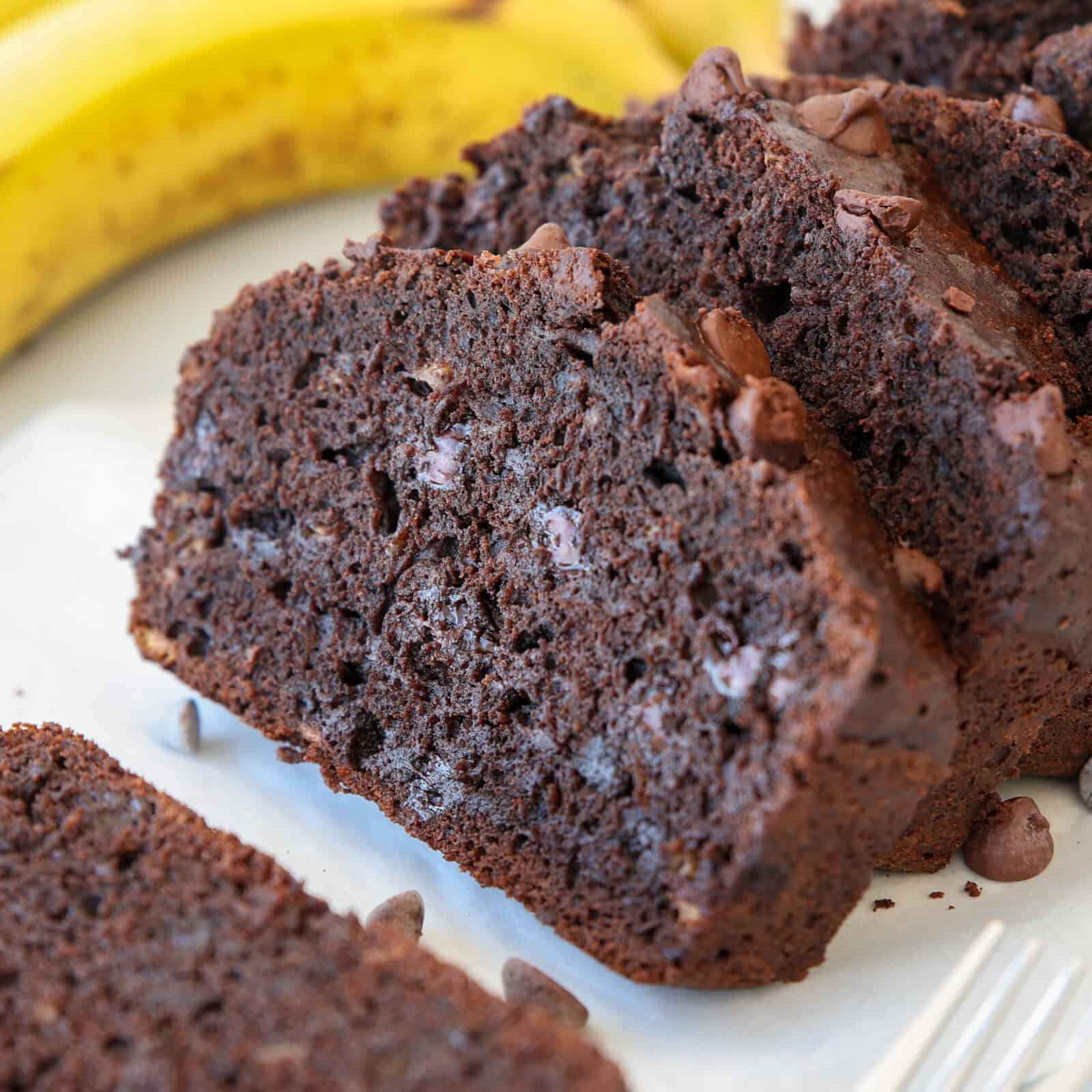 Close-up of sliced double chocolate banana bread with chocolate chips, arranged on a white surface, with a few ripe bananas in the background.