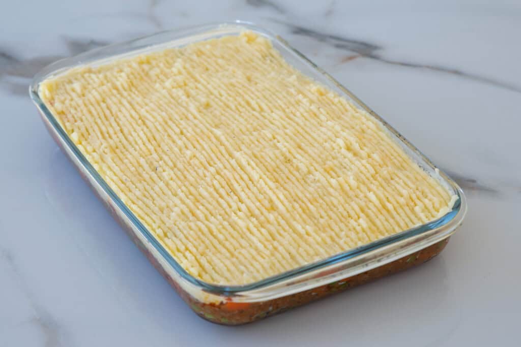 A rectangular glass baking dish filled with shepherd’s pie, showing a layer of mashed potatoes with a ridged pattern on top, set on a white marble surface.