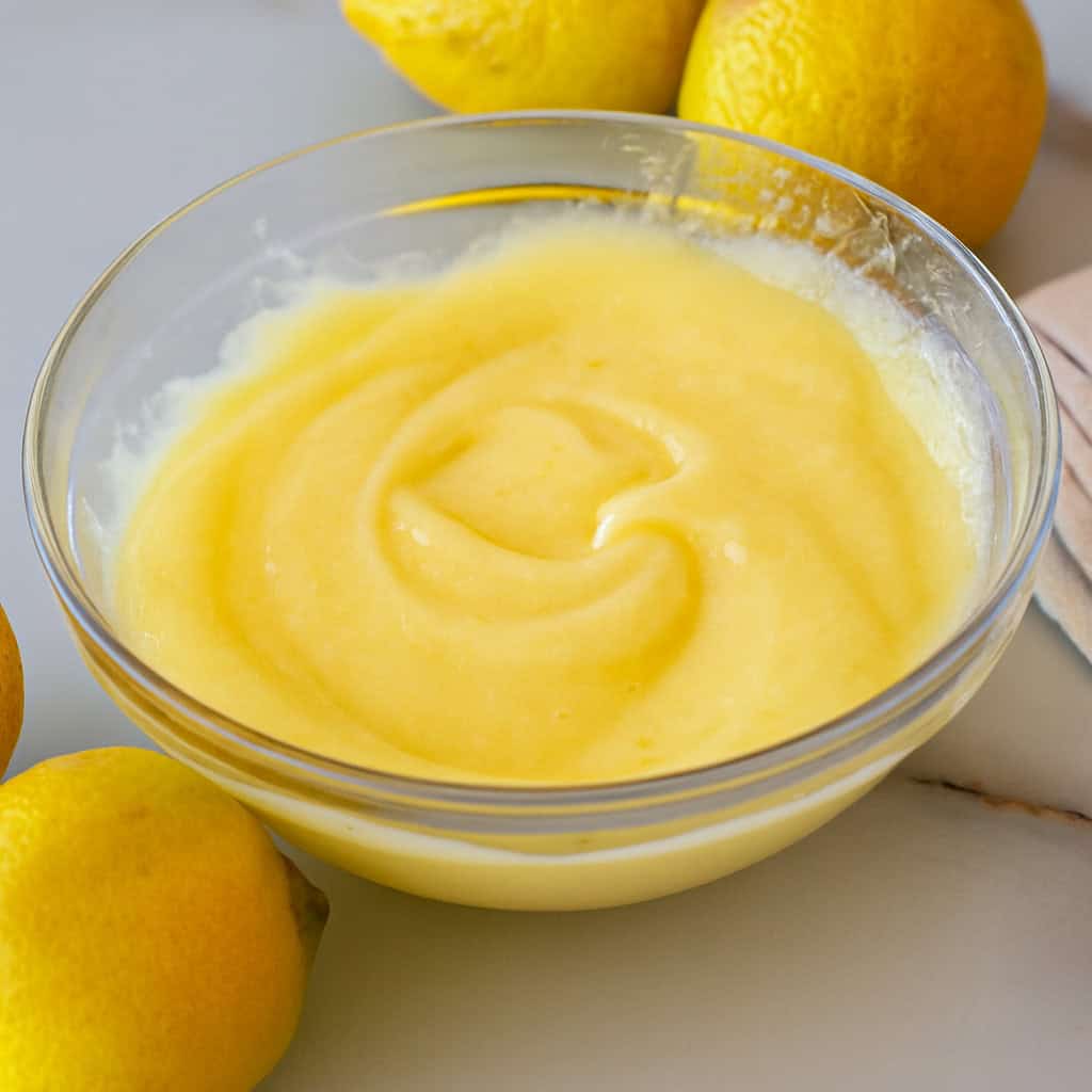 A clear glass bowl filled with smooth, creamy yellow lemon curd sits on a white surface, surrounded by whole lemons.