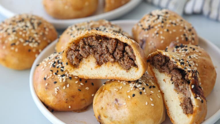 A plate of round, golden-brown meat buns topped with sesame seeds, some cut open to reveal a savory, seasoned ground beef filling inside.