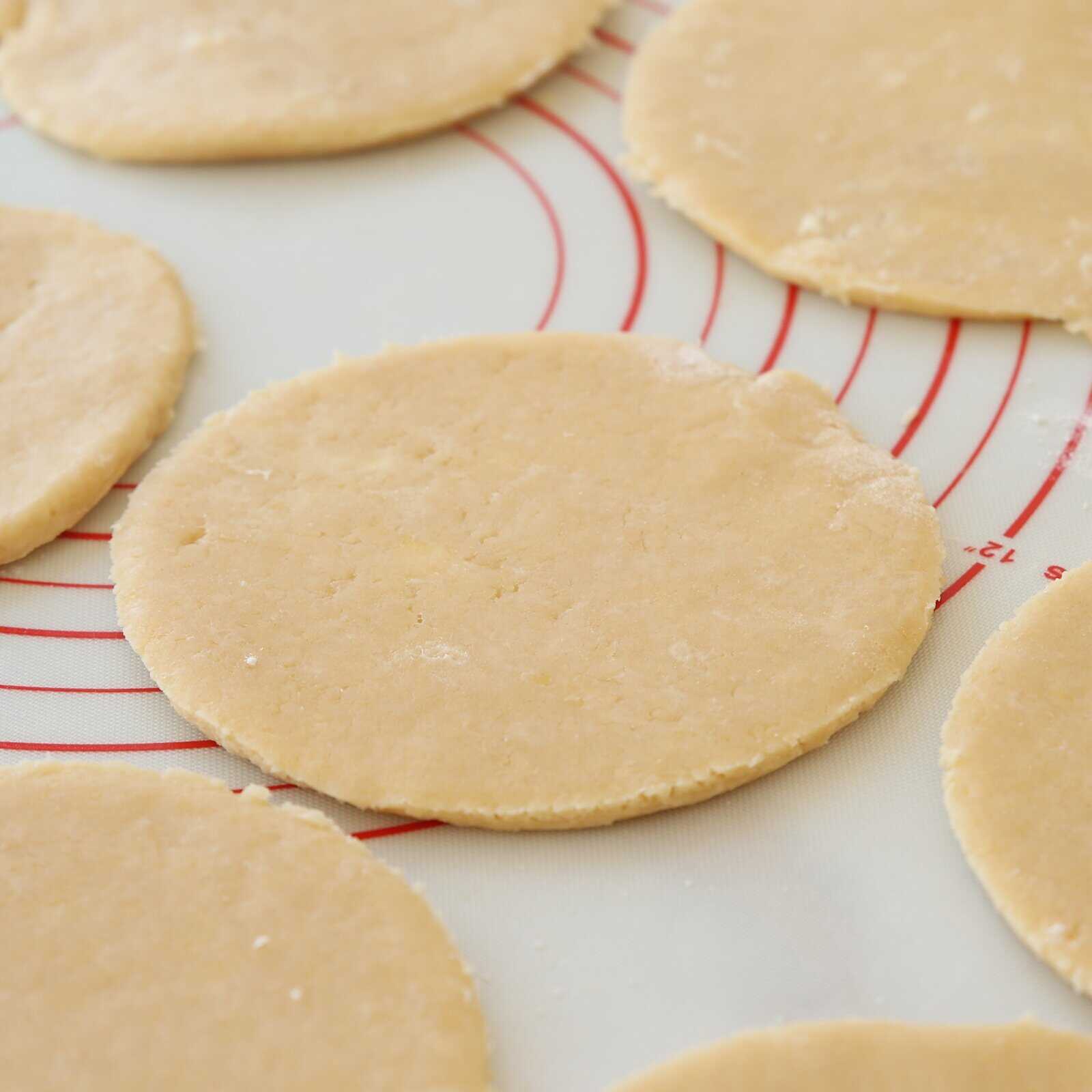 Several round, flattened pieces of raw dough are arranged on a silicone baking mat with red circular guides. The dough discs appear ready for baking or further preparation.