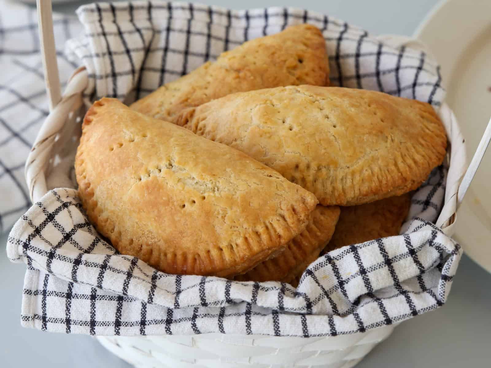 A white basket lined with a black-and-white checkered cloth holds several golden-brown, baked meat hand pies, showing a flaky crust and crimped edges.