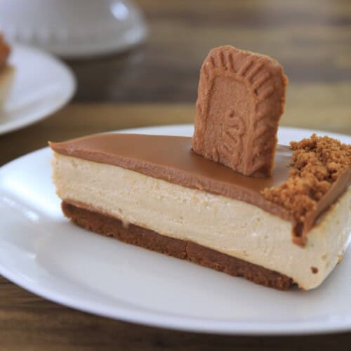 A slice of cheesecake with a Biscoff cookie butter topping and cookie base, garnished with a whole cookie and cookie crumbs, served on a white plate.