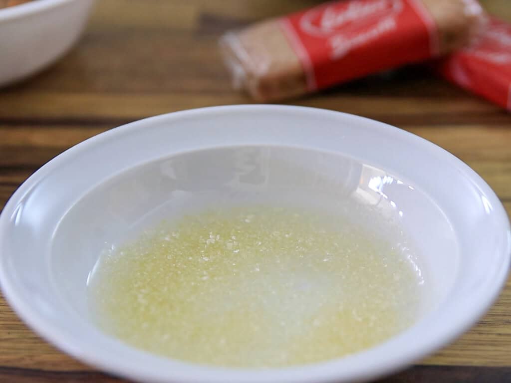 A white bowl contains a mixture of melted butter and sugar on a wooden surface. In the background, a packet of brown sugar and part of another white bowl are visible, slightly out of focus.
