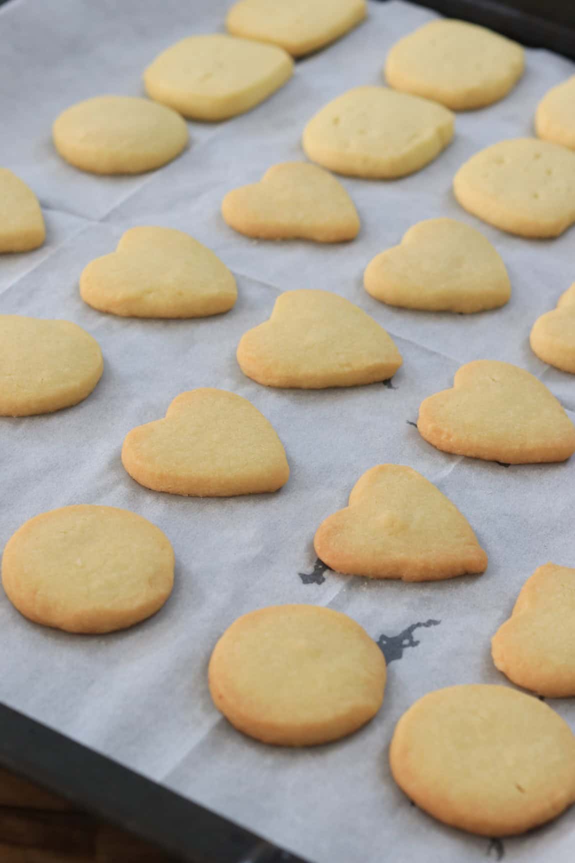 A baking tray lined with parchment paper holds rows of freshly baked shortbread cookies in heart and round shapes. The cookies are golden brown and evenly spaced.
