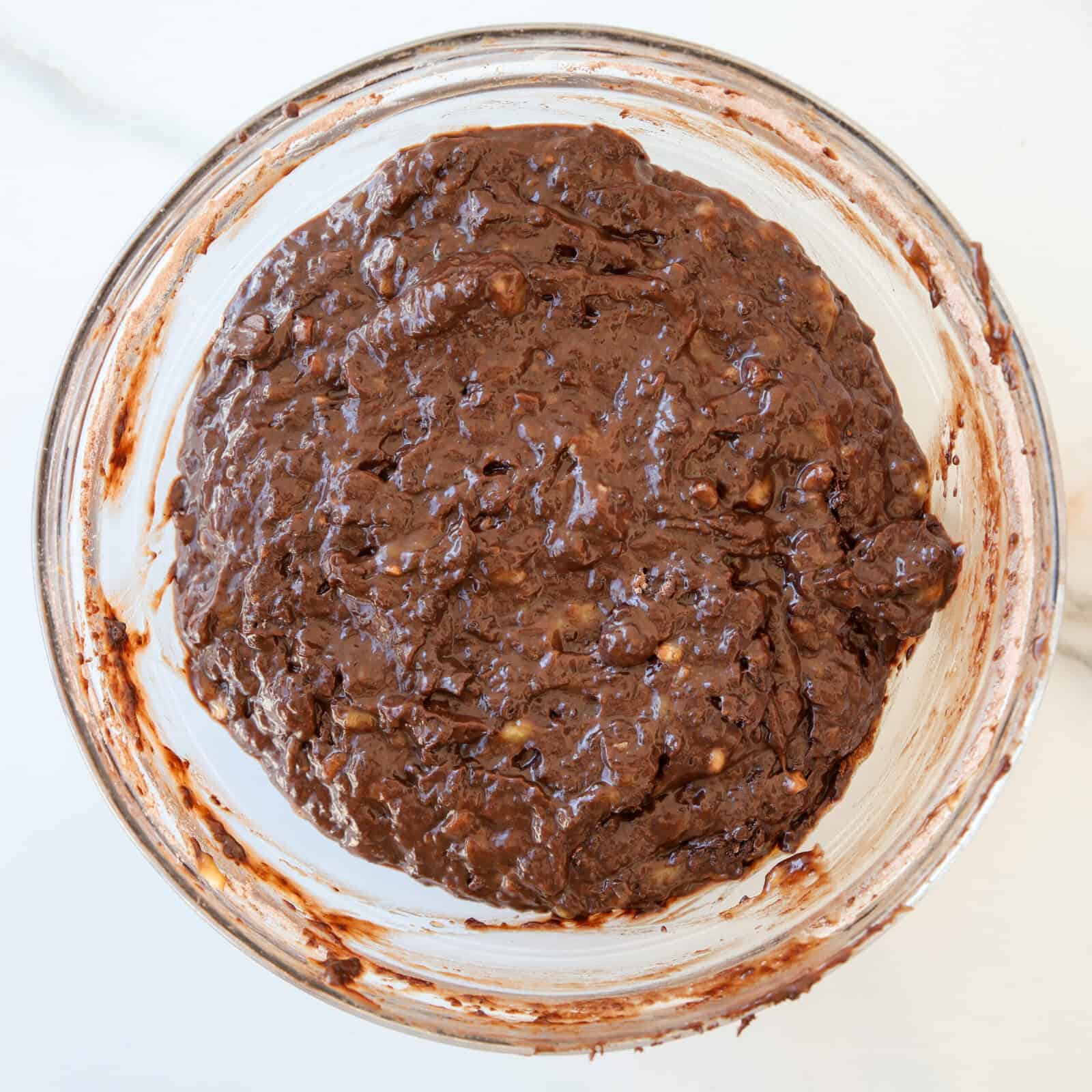 A glass bowl filled with thick, dark chocolate brownie batter mixed with chunks, sitting on a white surface.