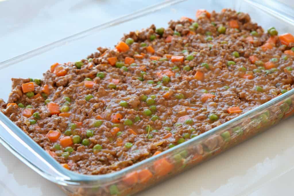 A glass baking dish filled with a mixture of cooked ground meat, green peas, and diced carrots in a brown sauce, evenly spread out and ready for the next step in meal preparation.