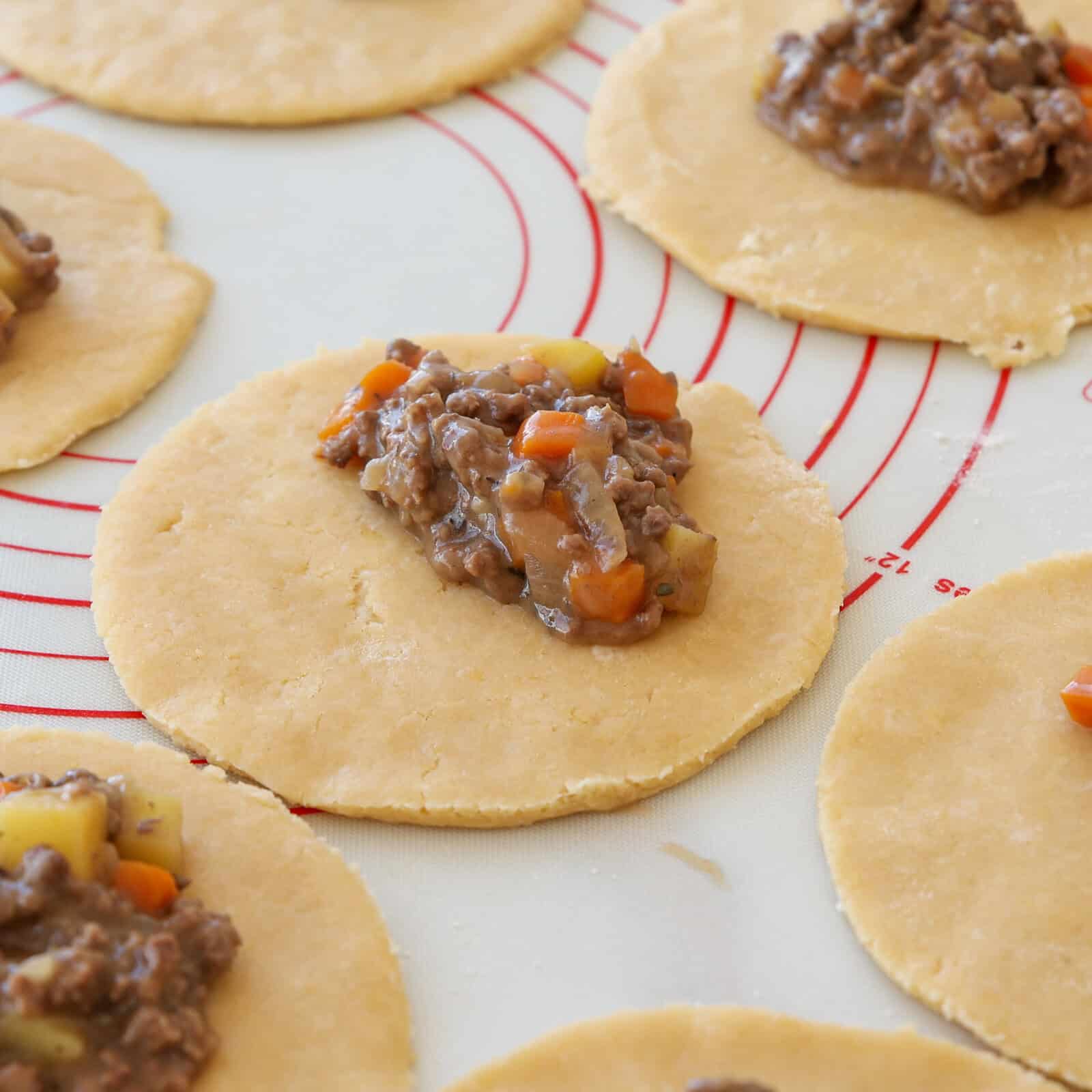 Round pastry dough circles with a scoop of ground meat, potato, and vegetable filling are arranged on a baking mat, ready to be folded and assembled, likely for making hand pies or turnovers.