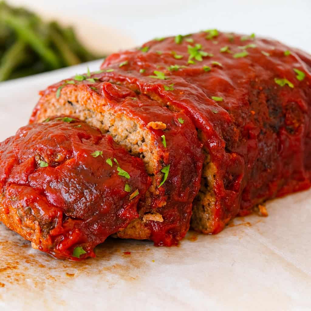 A freshly baked meatloaf topped with tomato sauce and chopped parsley is sliced and served on parchment paper. Green beans are blurred in the background.