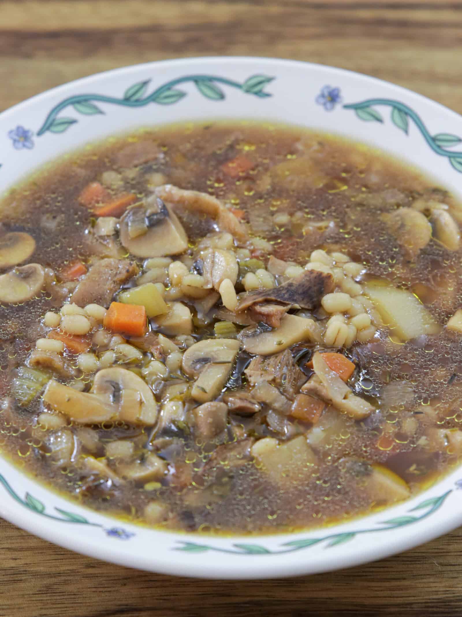 A bowl of vegan mushroom barley soup with visible mushrooms, barley, carrots, celery, and broth, served in a white bowl with a green and blue floral pattern on the rim, placed on a wooden surface.