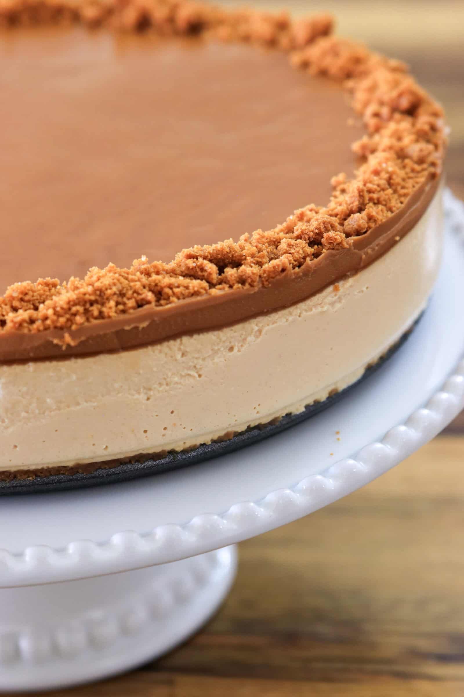 A creamy cheesecake with a light brown, smooth topping and a crumbly cookie crust around the edge sits on a white cake stand with a beaded rim.