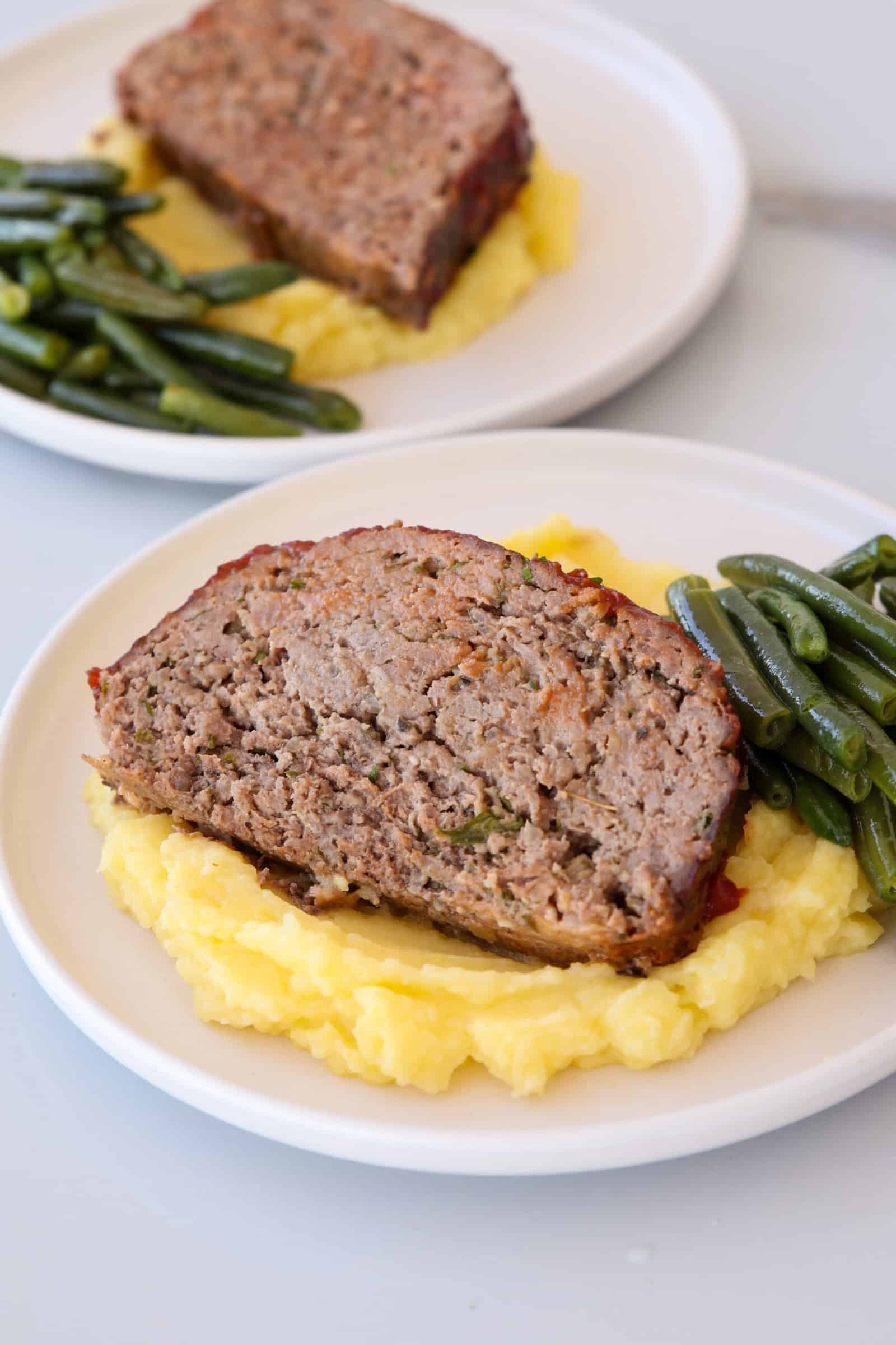 A slice of meatloaf served on mashed potatoes with a side of green beans on a white plate. Another similar plate is visible in the background.