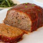A close-up of a sliced meatloaf topped with a tomato glaze and sprinkled with chopped parsley, resting on parchment paper. A plate of green beans is blurred in the background.