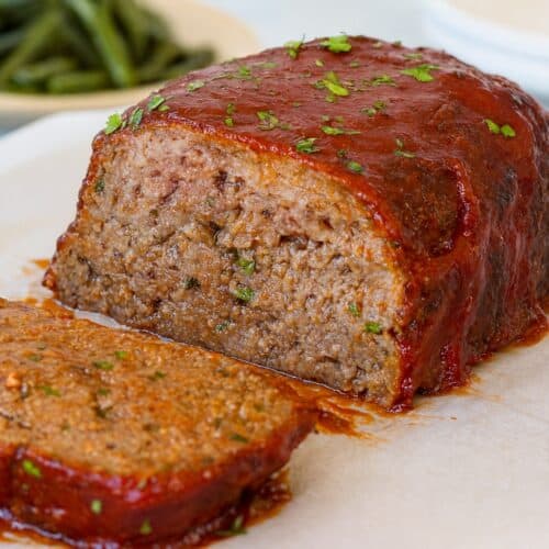 A close-up of a sliced meatloaf topped with a tomato glaze and sprinkled with chopped parsley, resting on parchment paper. A plate of green beans is blurred in the background.