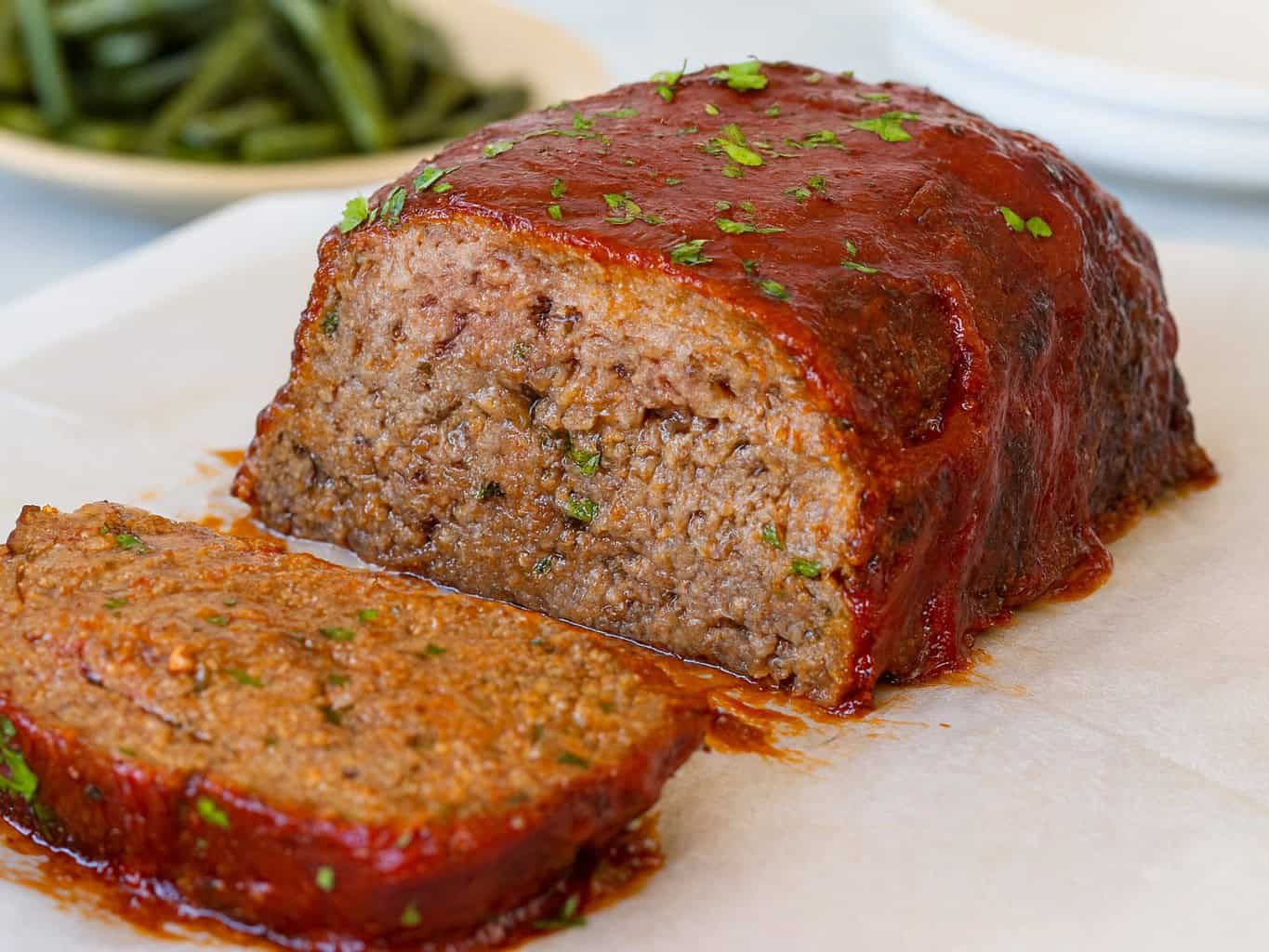 A close-up of a sliced juicy meatloaf topped with a tomato glaze and sprinkled with chopped parsley, resting on parchment paper. A plate of green beans is blurred in the background.