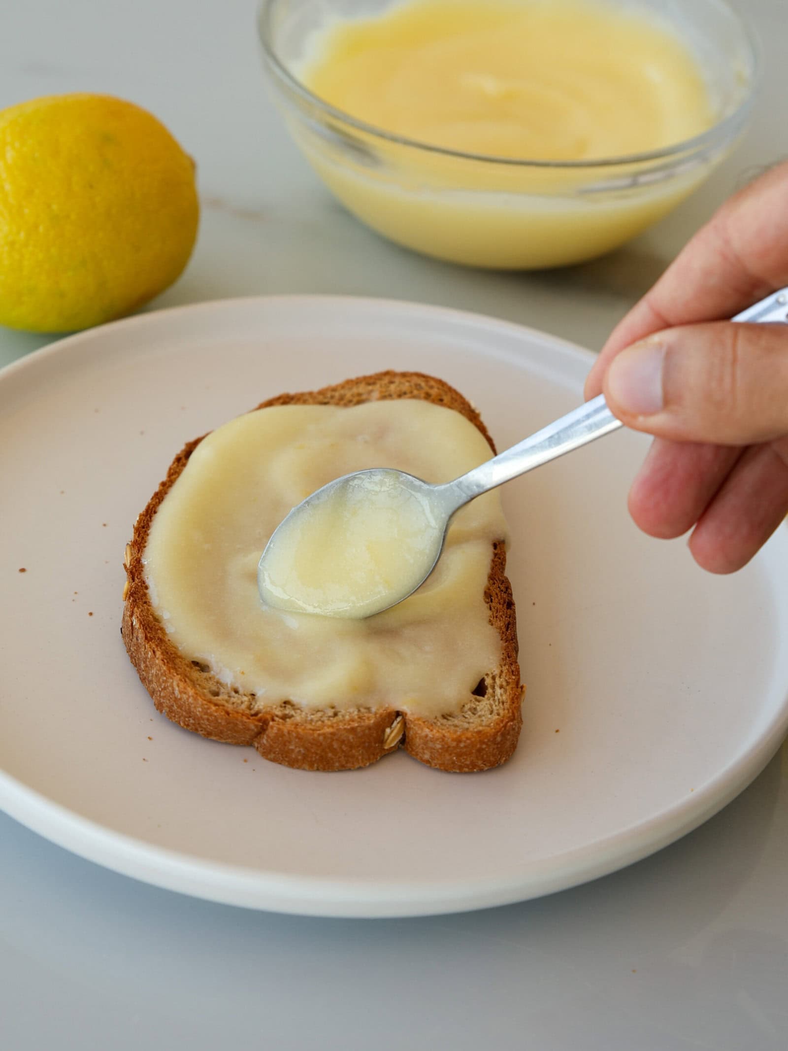 A hand spreads homemade lemon curd onto a slice of bread with a spoon. A lemon and a bowl of lemon curd are in the background on a light surface.