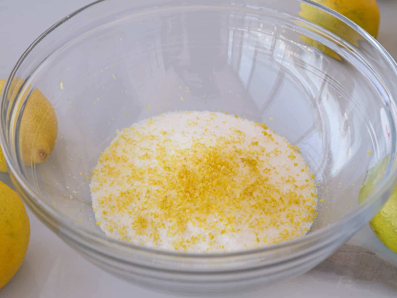 A clear glass bowl containing white granulated sugar topped with fresh lemon zest. Whole lemons are visible around the bowl on a light-colored surface.