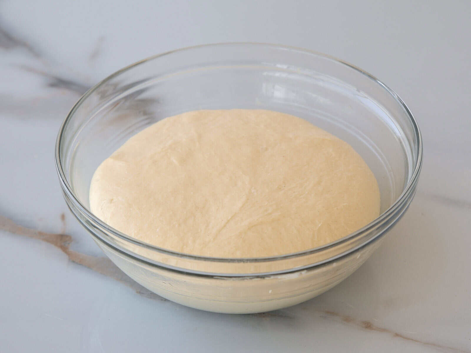 A glass bowl with risen bread dough resting on a light marble surface. The dough appears smooth and has expanded, filling most of the bowl.