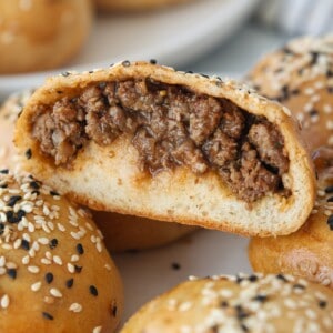 A close-up of a baked bread roll filled with seasoned ground beef, topped with white and black sesame seeds. One roll is cut in half to show the savory meat filling inside. Other rolls are visible in the background.