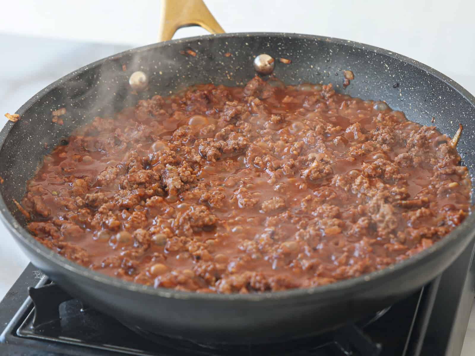 A frying pan filled with ground beef and tomato sauce mixture simmering on a stovetop, with steam rising from the food.