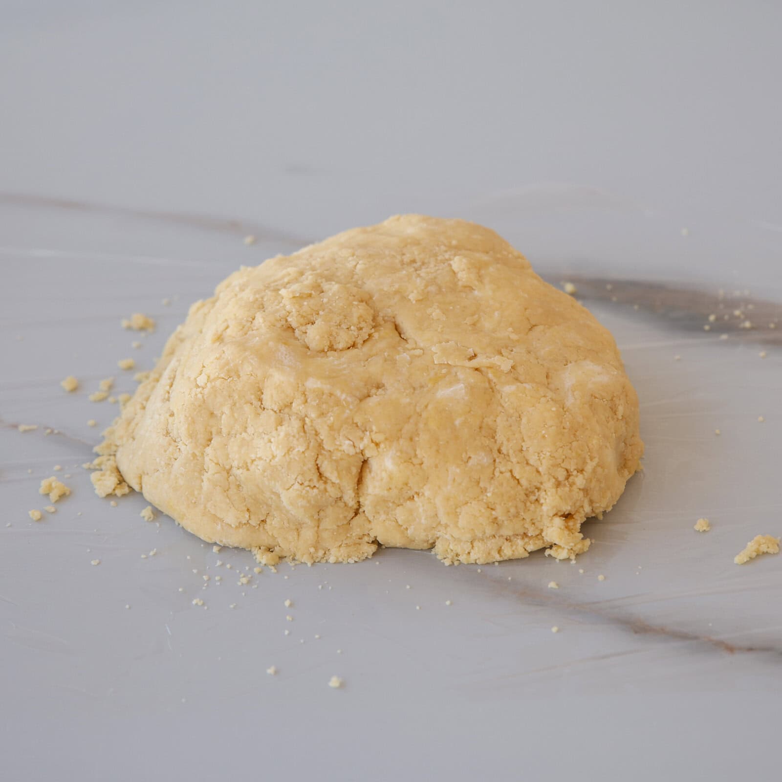 A mound of rough, yellowish dough sits on a smooth, light-colored surface with a few small crumbs scattered around it.