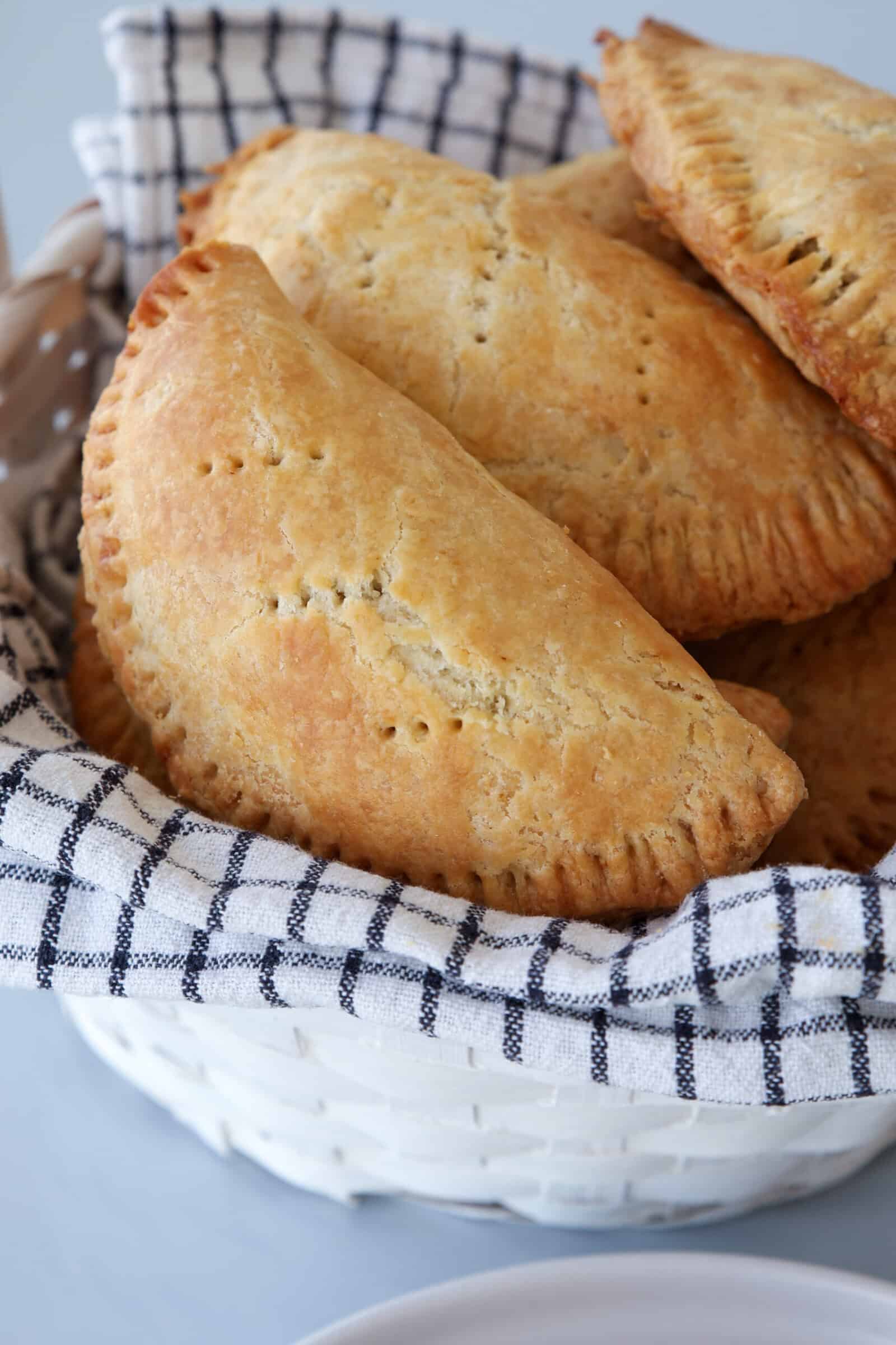 A basket lined with a black-and-white checkered cloth holds several golden-brown, baked Nigerian meat pies with crimped edges.