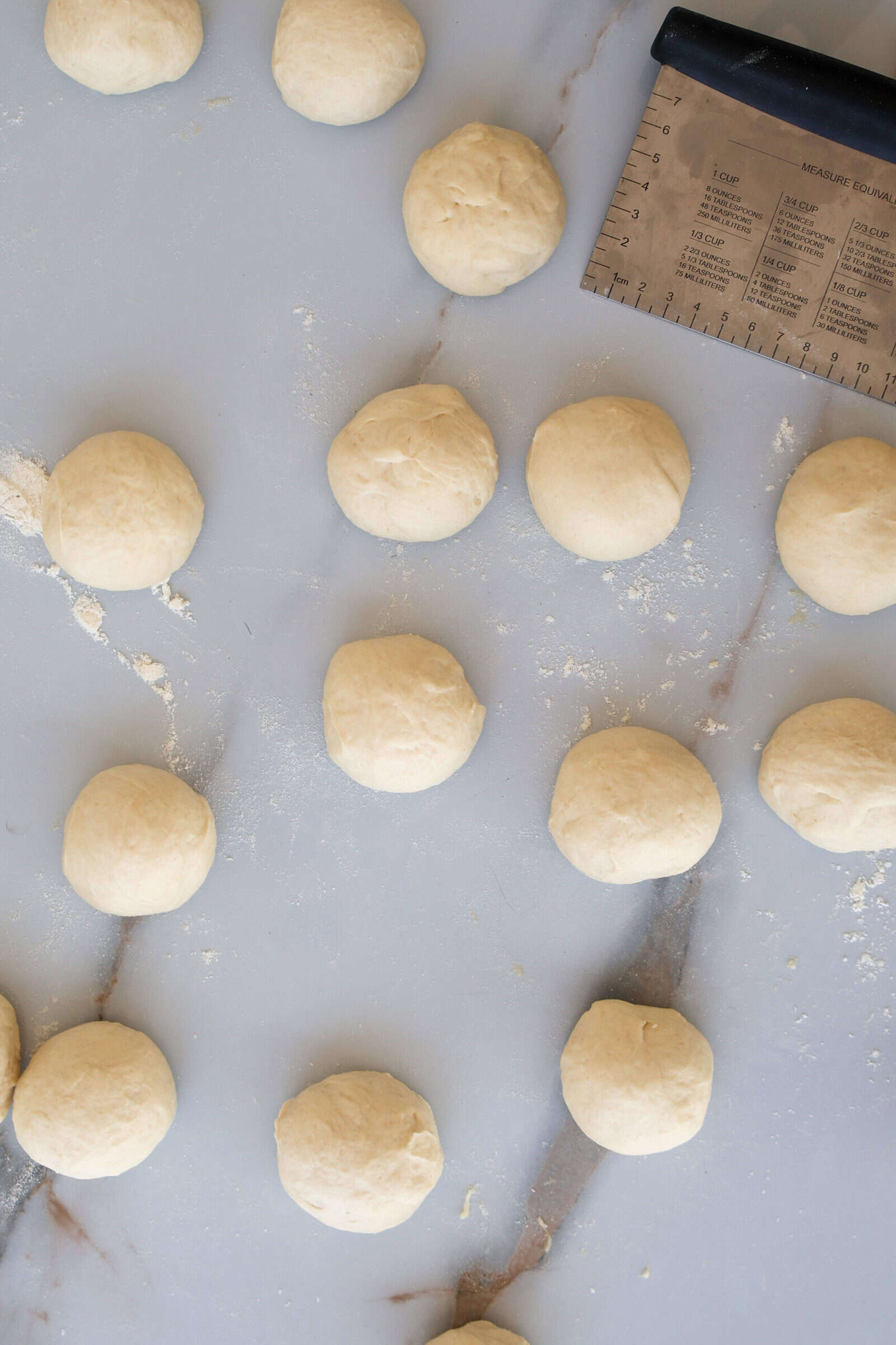 Several evenly spaced balls of dough rest on a lightly floured marble surface, with a metal bench scraper featuring measurement markings visible in the upper right corner.